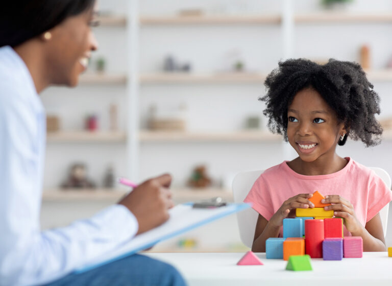 Friendly attractive african american woman child development specialist having session with cheerful little girl, sitting at table, playing with colorful wooden bricks, having conversation and smiling